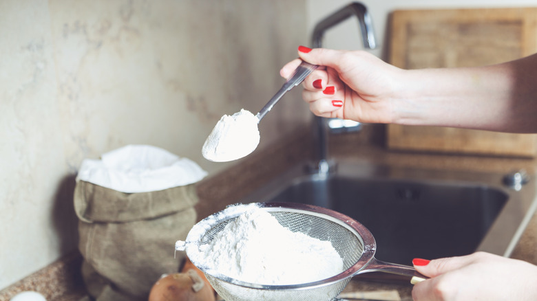 Someone scooping flour with a spoon from a sifter near a sink
