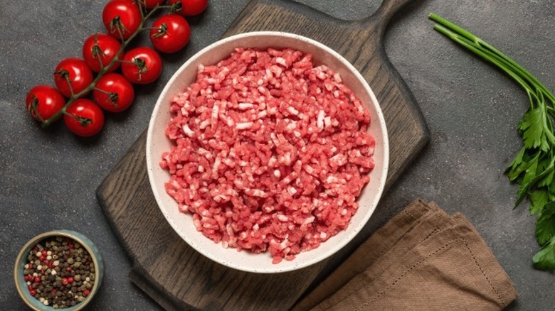 Raw ground beef in a bowl on a black surface with pepper in a ramekin and cherry tomatoes on the vine nearby.