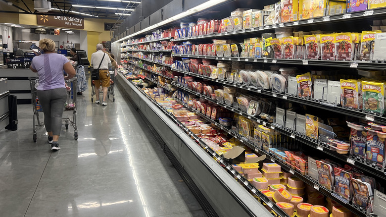 Grocery shoppers push their carts past a wall of packaged deli meat