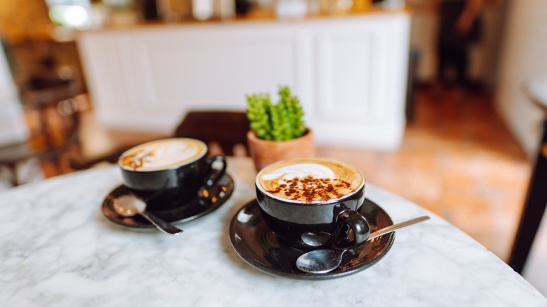 Two latte drinks sitting on a cafe table with cafe interior blurred in background