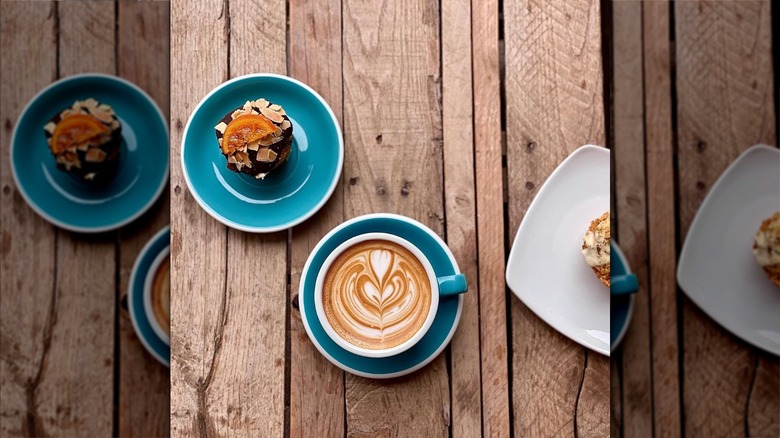 Muffin topped with an orange slice on a plate beside latte with latte art on a wooden table, view from above, at Iconik Coffee Roasters