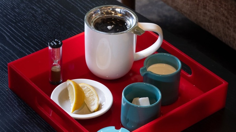 Coffee mug with side mugs of cream and sugar cubes and a timer and sliced lemon all on a red tray at Red Rooster Coffee