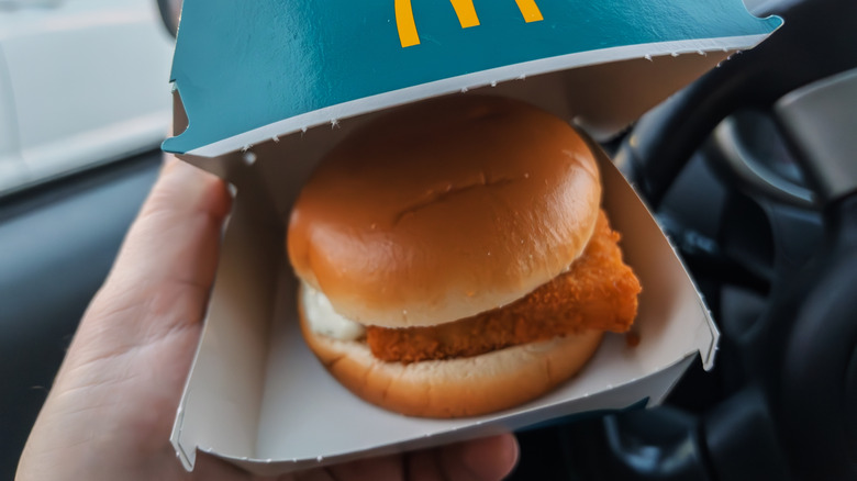 The hand of a person sitting behind the wheel of a car holds a Filet-O-Fish container