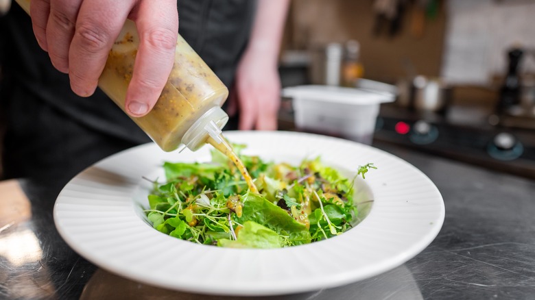 Person using squeeze bottle filled with grainy salad dressing to drizzle onto white plate of delicate greens