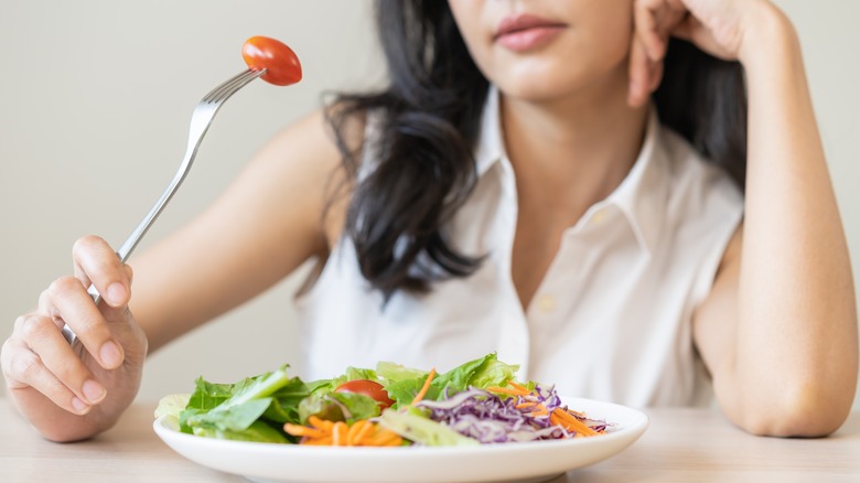 girl looking reluctantly at cherry tomato on. fork held above mixed salad