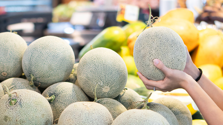 A woman's hands lift a cantaloupe from a supermarket stack