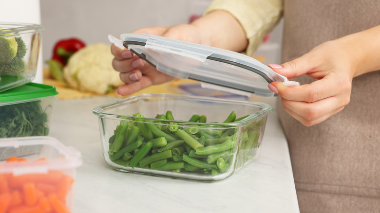 A person putting the lid on a glass food storage container full of cooked green beans.