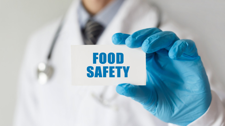 A man in a white lab coat and blue latex gloves holding a placard reading "food safety"