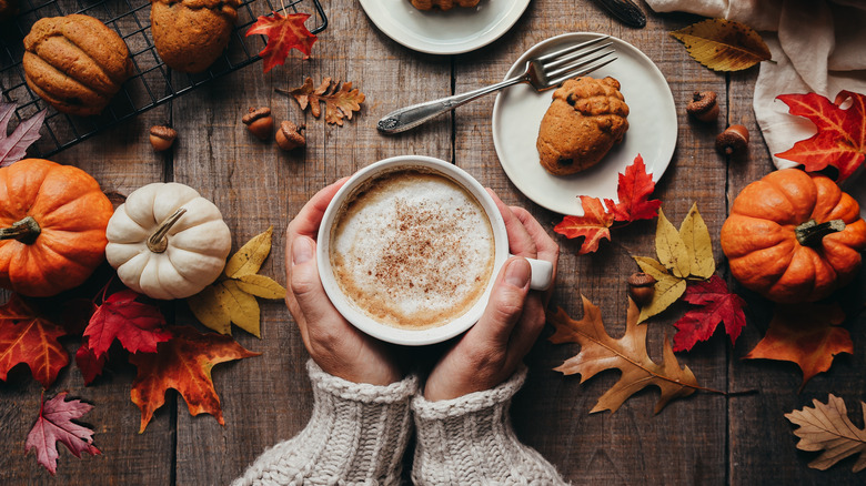 Hands holding a mug with pumpkin spice coffee surrounded by small pumpkins, fall leaves, and an acorn-shaped small cake