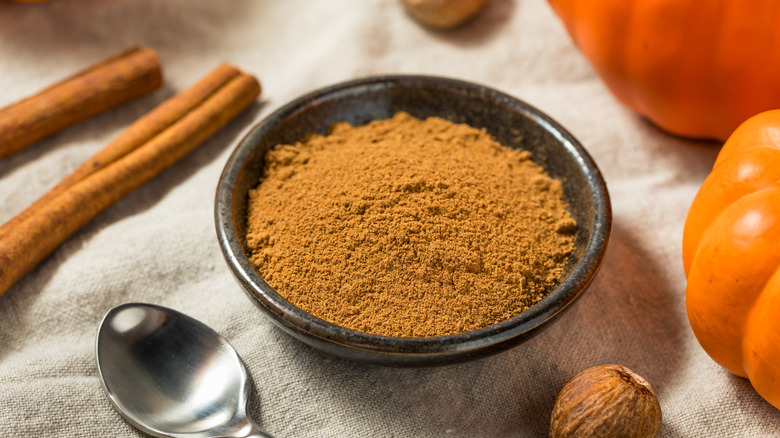 Small bowl of pumpkin spice alongside small pumpkins, cinnamon sticks, and nutmeg