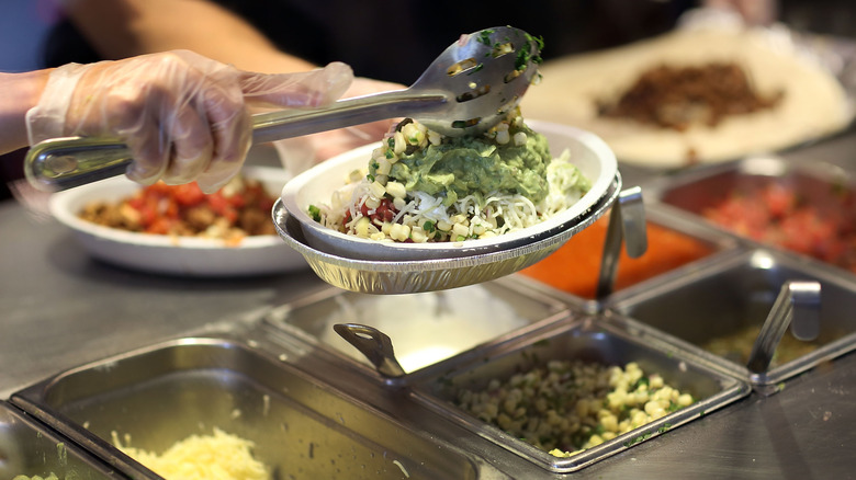 Hands of Chipotle employee with spoon piling toppings onto burrito bowl