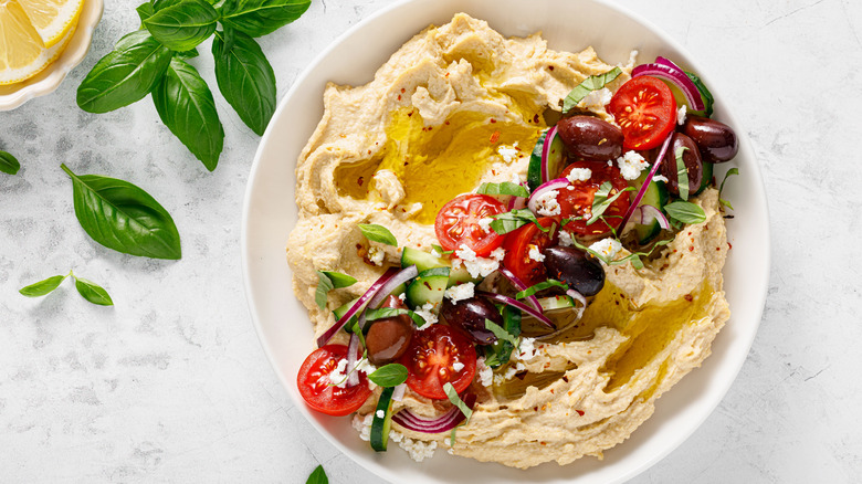 A bowl of hummus with tomatoes, onions, olives, cucumbers, herbs, and feta