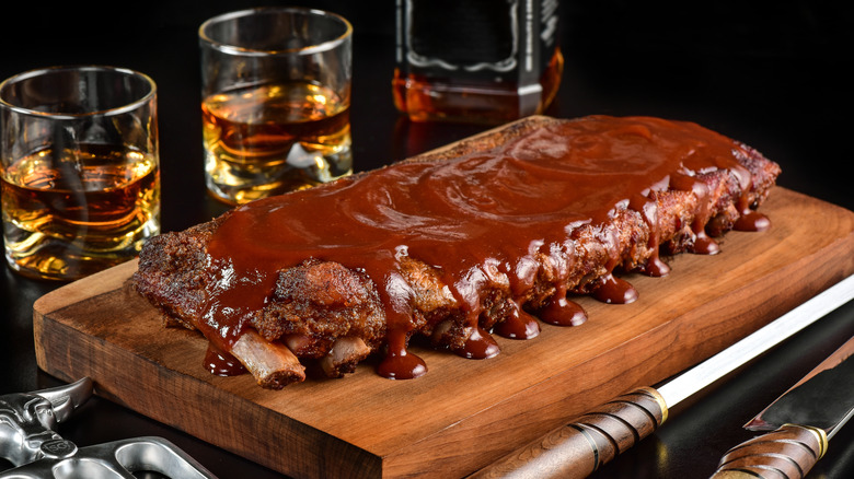 Glistening BBQ ribs on wood board with lots of sauce on it and whiskey in the background.