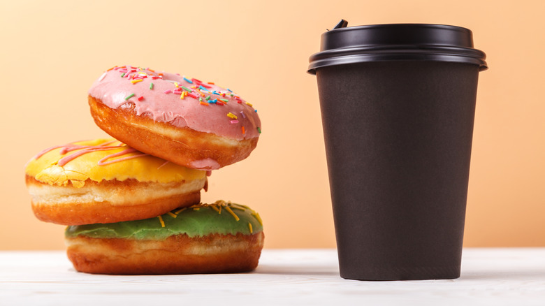 Three donuts frosted pink, yellow, and green stacked next to a dark brown coffee cup with a black lid