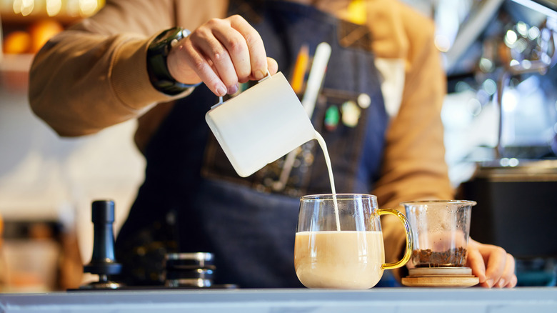 Person wearing apron and watch pouring cup of milk into a cup of tea