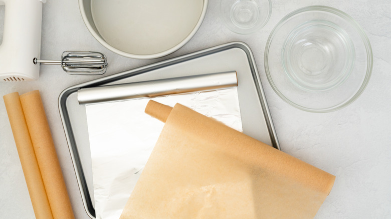 Parchment paper and aluminum foil rolls lay partially unrolled on a baking pan. Glass bowls, a pan, a mixer and more rolled paper is strewn around the pan.