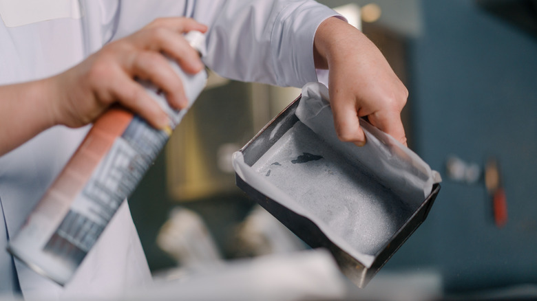 A baking pan with parchment paper is held in one hand, while cooking spray is in the other. The pan is being sprayed, with a blurred background.