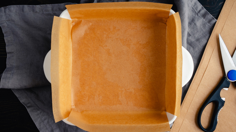 A square baking pan is filled with parchment paper that is folded over the sides. In the background is a kitchen cloth and scissors.