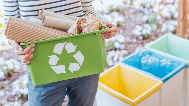A person holds a green recycling bin stacked high with cardboard and paper. In the background there are yellow, blue and green bins, and some greenery.