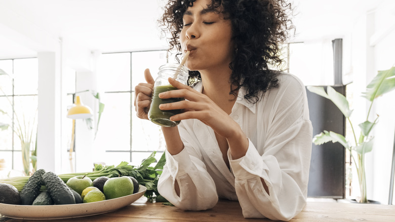 A woman sips a green smoothie through a straw next to a plate of vegetables