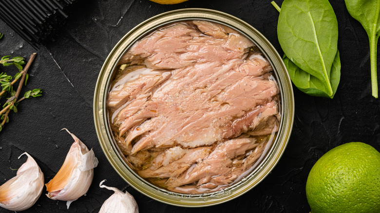Top view of an open can of tuna surrounded by garlic cloves, spinach leaves, thyme, and whole lime