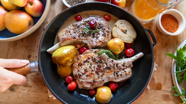 Pok chops, apples, cherries, and rosemary in black skillet with fresh apples and cider nearby