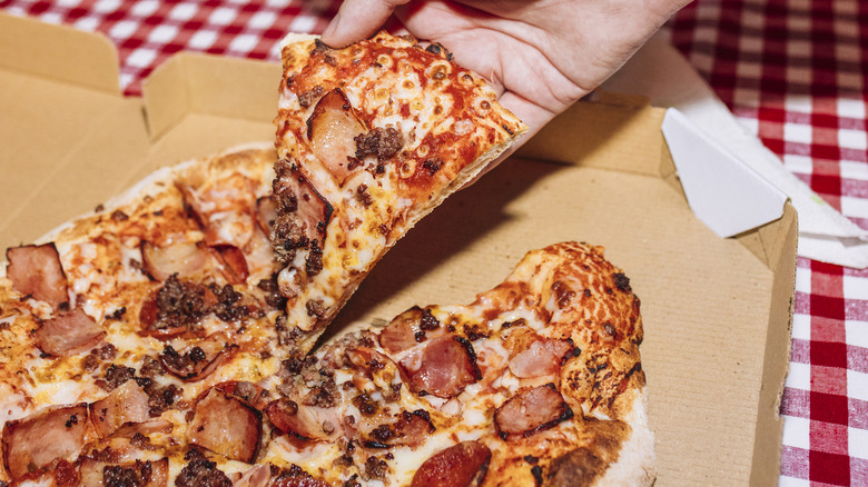 A hand takes a slice from a pizza topped with meats