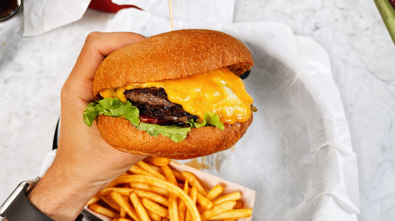 A person's hand raises a cheeseburger over a container of fries.