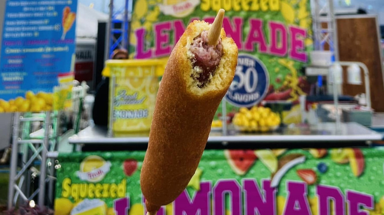 A Conecuh corn dog with a bite taken out of it, held in front of a fair concession stand.
