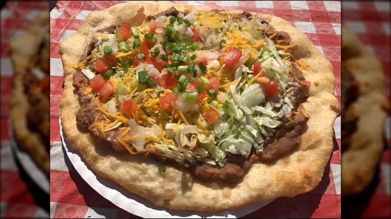 A Navajo Taco: fry bread topped with beans, shredded cheese and vegetables on checkered cloth.
