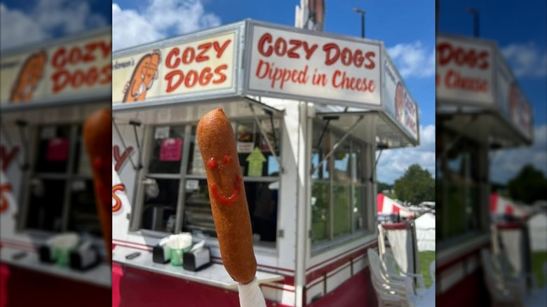 A Cozy Dog with a ketchup-drawn smiley face is held up in front of a Coleman's Concession Cozy Dog stand.