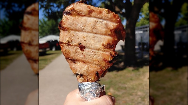 Grilled pork chop, bottom wrapped in foil, held up with blurred outdoor background.