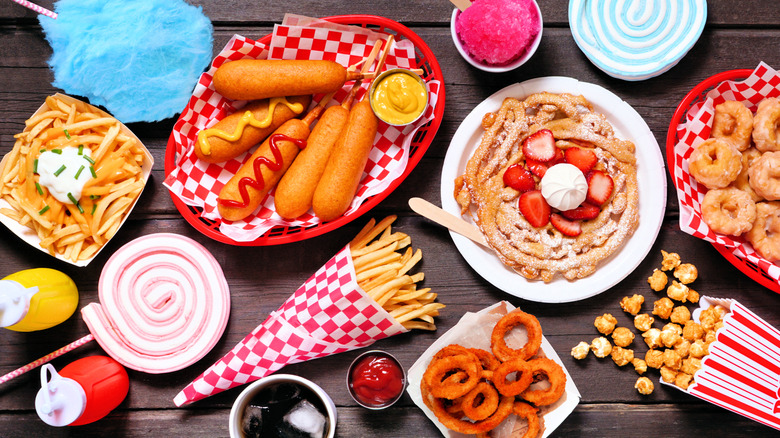 A table full of fairground treats including, cotton candy, fries, corndogs, snow cone, funnel cake, mini donuts, onion rings, caramel corn, and soda.