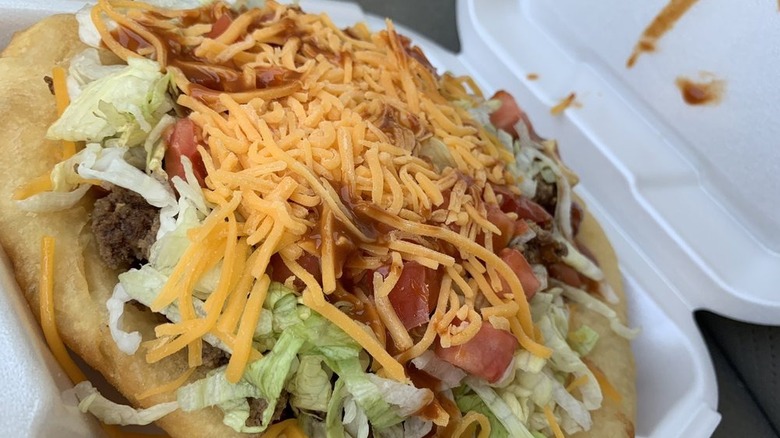 A close up of fry bread taco covered with meat, lettuce, tomatoes, cheese and hot sauce.