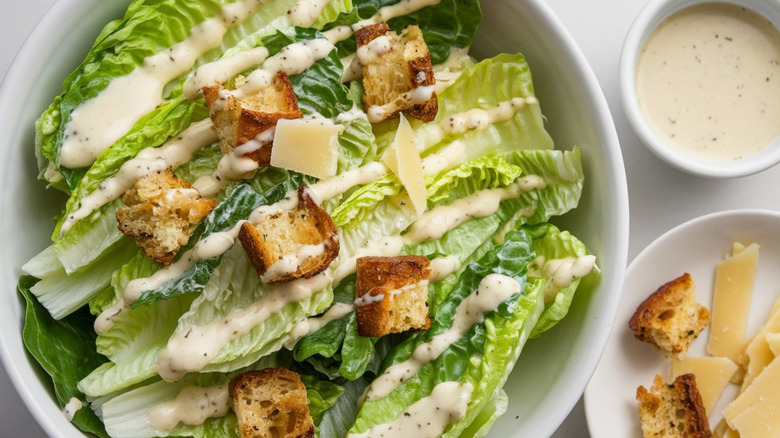 Caesar salad in white bowl with a small ramekin of dressing on the side. Croutons and parmesan are on a side plate nearby.