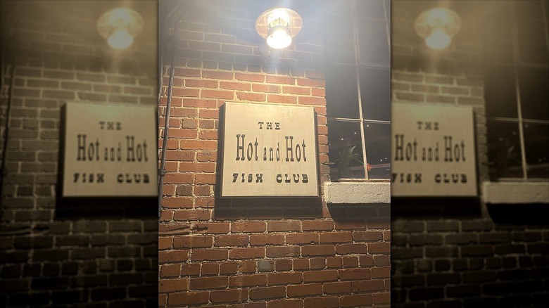 Exterior of a brick building with a sign lit by a single light reading "The Hot and Hot Fish Club"