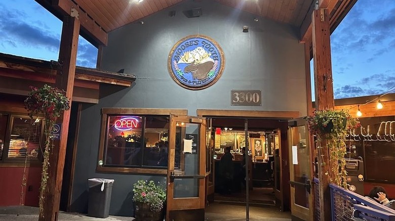 Entrance of Moose's Tooth Pub & Pizzeria at dusk; large wooden gable over the door, a circular logo with a moose illustration on gray painted exterior