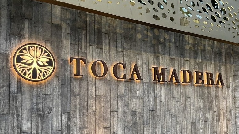 The weathered wood exterior of Toca Madera restaurant; a round metal backlit flower/tree logo on the left and backlit metal letters spelling "Toca Madera" on the right