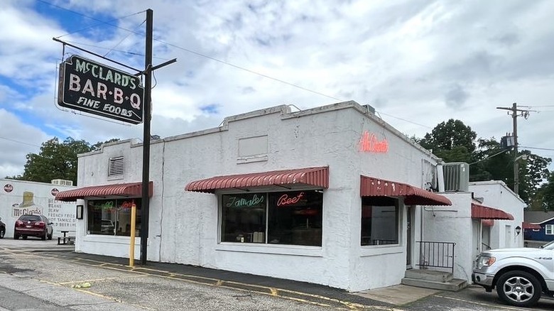 The exterior of the McClard's Bar-B-Q building; weathered white concrete single-story  building with two large windows on one side and one on the other. windows have red awnings; vintage black neon sign on the left reads "McClard's Bar-B-Q Fine Foods"