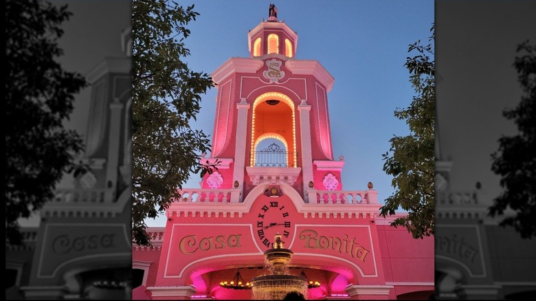 Bright pink tower exterior of Casa Bonita at dusk with a gold fountain in the foreground; building is trimmed in white; "Casa Bonita" appears in gold letters, surrounding a clock