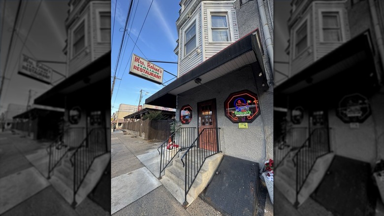 The exterior entrance of Mrs. Robino's restaurant; old building with gray stucco on first floor and gray siding and windows on the second floor; awning appears over concrete steps to door; white vintage "Mrs. Robino's Italian Restaurant" hangs on the left