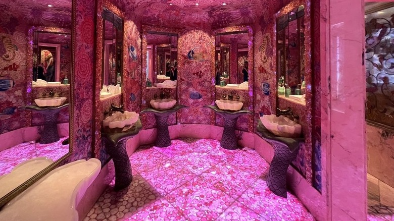 Bathroom at Sexy Fish lit in bubblegum pink; opulent alcove with four shell-shaped sinks, pebble-tiled floor, marble walls and elaborate wallpaper and mirrors on walls