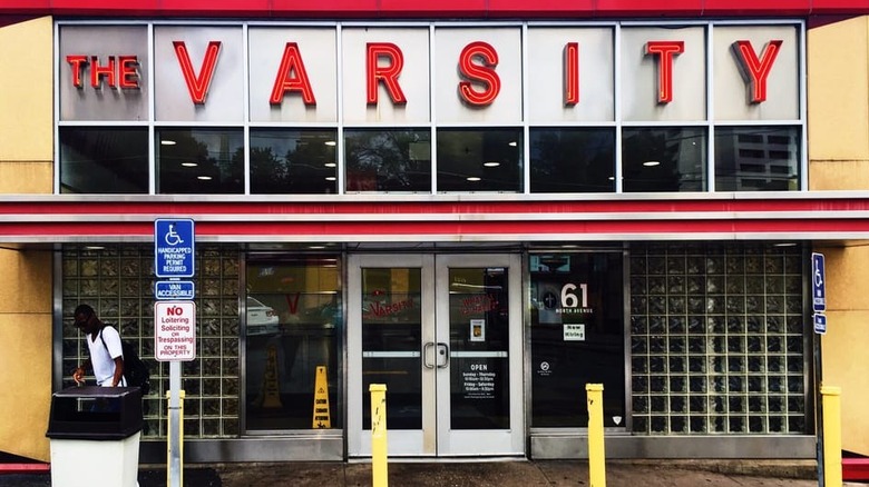 Exterior of The Varsity; standard business double-doors flanked by windows and glass bricks; "The Varsity" is spelled out over a red stripe border in red on a white background; exterior walls are yellow