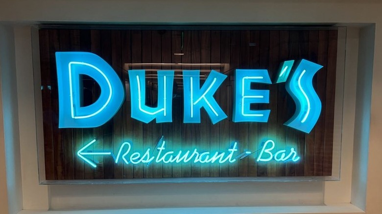 A window with a neon sign that reads "Duke's" in bright blue neon letters with "Restaurant - bar" in blue neon underneath and an arrow pointing left