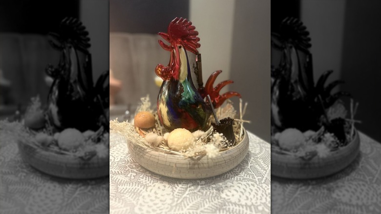 A gray shallow bowl set on a gray pinecone-patterned tablecloth with nest- and egg-like food sculptures in it with a food sculpture of a rooster sitting in the center