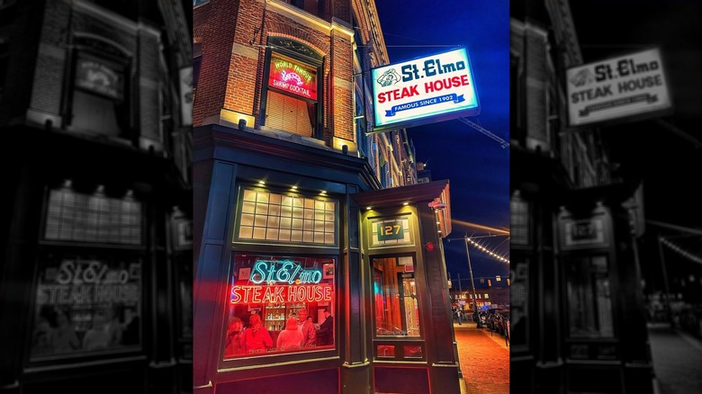 Exterior of St. Elmo Steak House at night; two-story brick building with a blue and red neon sign in left window reading "St. Elmo Steak House" and a neon sign in the window abovie it and a sign above the door reading "St. Elmo Steak House" in blue and red letters on a white background