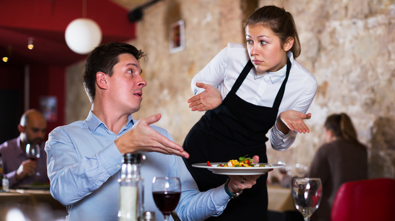 A man, left, seated at a table in a restaurant, holding a plate of food and gesturing toward it unhappily as a young female server shrugs her shoulders as if she doesn't care