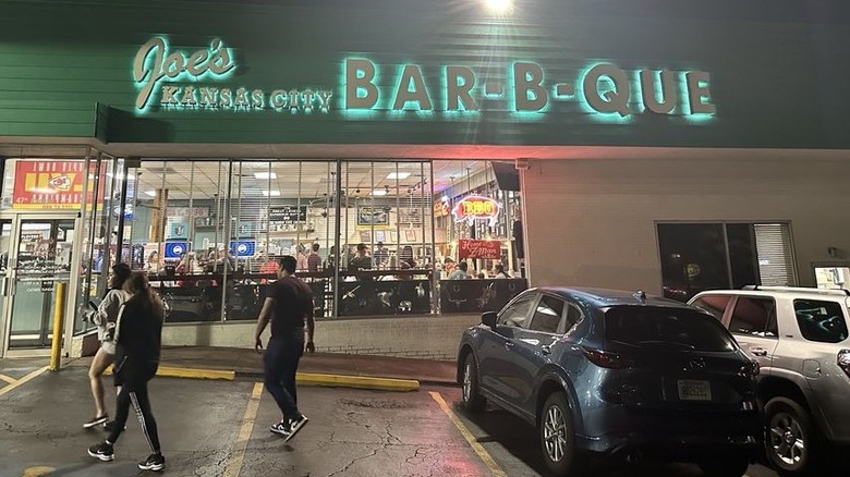 The exterior of a convenience store from the parking lot at night; a green backlit sign on a greeen background reads "Joe's Kansas City Bar-B-Que"