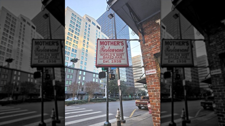 A corner on an urban street showing a white vintage sign reading, "Mother's Restaurant World's Best Baked Ham Est. 1938" in read letters affixed to a brick building
