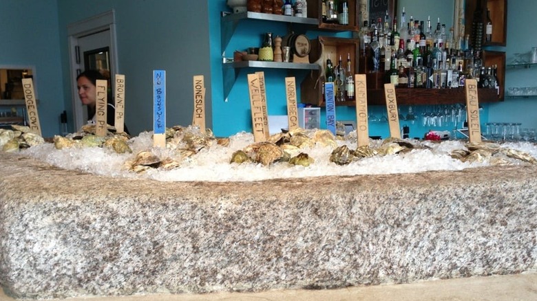 Interior of Eventide restaurant; large rock container containing ice and oysters labeled with beige and blue sticks; liquor bottles in the background